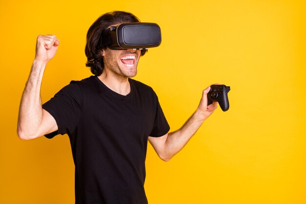 Photo de jeune homme réalité virtuelle tenir joystick bouche ouverte lever le bras poing porter casque vr t-shirt isolé fond de couleur jaune