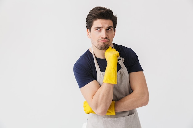 Photo d'un jeune homme séduisant portant des gants en caoutchouc jaune pour la protection des mains posant pendant le nettoyage de la maison isolée sur blanc