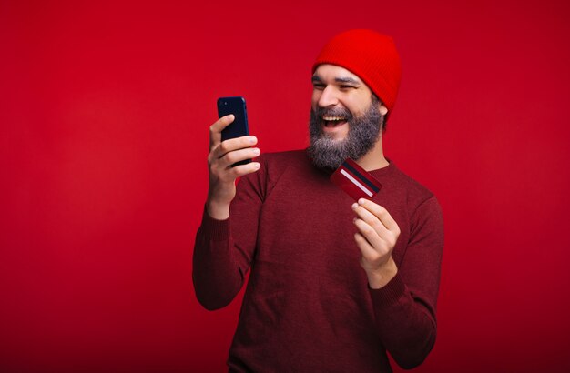 Photo de jeune homme souriant avec barbe blanche regardant smartphone et tenant la carte de crédit, concept bancaire web