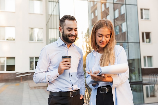 Photo d'un jeune homme souriant positif et d'une femme d'affaires à l'extérieur dans la rue près du centre d'affaires tenant un presse-papiers avec des plans de documents et une tasse de café.