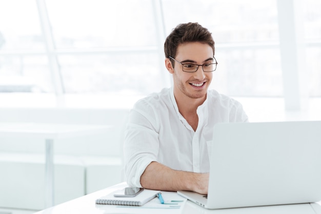 Photo d'un jeune homme souriant vêtu d'une chemise blanche à l'aide d'un ordinateur portable. Regardez un ordinateur portable.