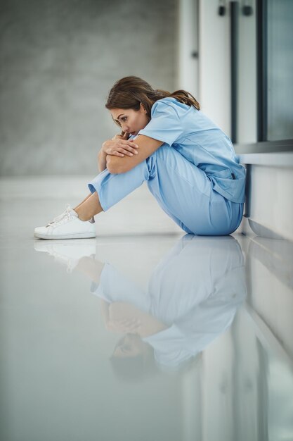 Photo d'une jeune infirmière fatiguée assise sur le sol près d'une fenêtre tout en faisant une pause rapide dans un couloir vide de l'hôpital pendant la pandémie de Covid-19.