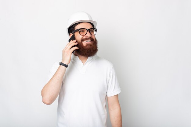 Une photo d'un jeune ingénieur barbu parlant à son téléphone tout en souriant et debout près d'un mur blanc