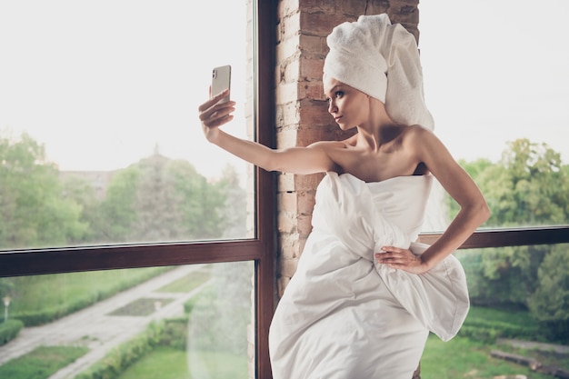 Photo d'une jeune jolie dame relaxant les procédures de la maison du spa quarantaine rester à la maison épaules nues tenir le téléphone faire des selfies blogger porter seulement une couverture tête turban grande fenêtre salon à l'intérieur