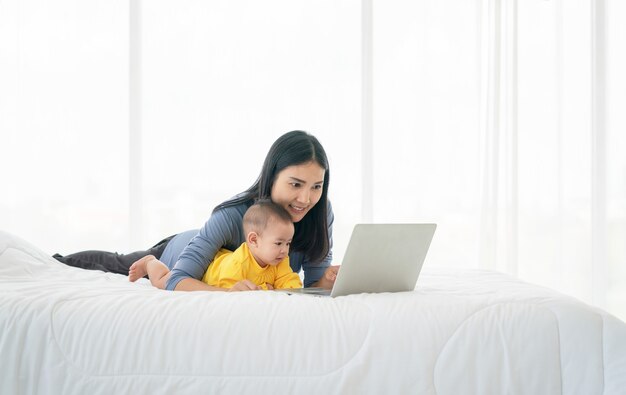 Photo de jeune maman thaïlandaise heureuse avec son bébé à l'aide d'un ordinateur portable sur le lit. Confort de la maison. Soin et attention. Travail à domicile.