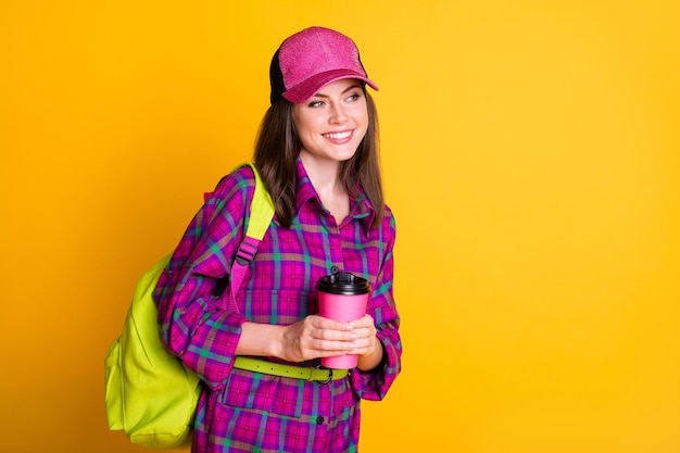 Photo d'une jolie adolescente joyeuse tenir la tasse aller à l'école porter un chapeau rose chemise verte sac ceinture isolé sur fond de couleur jaune