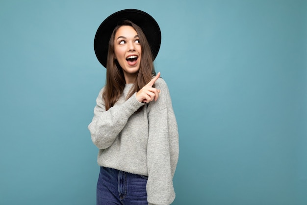 Photo d'une jolie brune heureuse choquée joyeuse jeune femme pointant le doigt vers l'espace libre pour le texte portant un maillot gris et un chapeau noir isolé sur fond bleu.