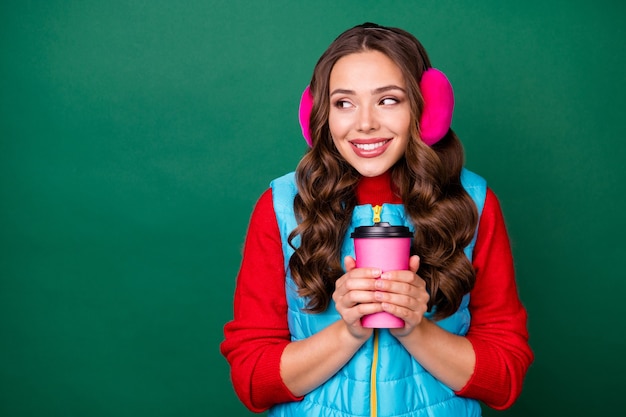 Photo de jolie charmante jeune femme tenir une tasse de café à emporter regarder un espace vide rayonnant souriant parc d'hiver marcher porter des cache-oreilles roses gilet bleu pull rouge isolé fond de couleur verte