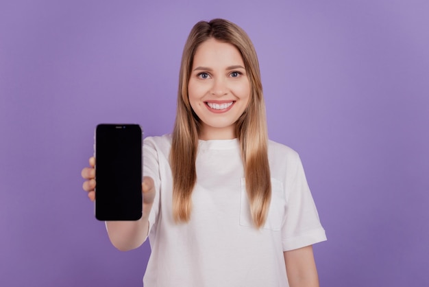 Photo d'une jolie dame assez positive tenir le téléphone montrer un espace vide à écran tactile porter un t-shirt décontracté sur fond violet