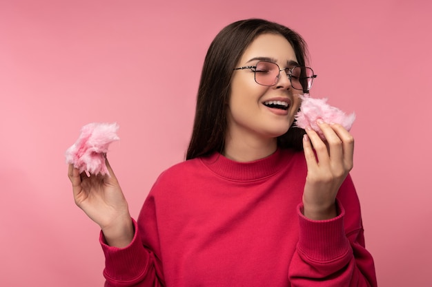 Photo de jolie dame dans des verres joue avec des bonbons de barbe à papa a du plaisir à porter un pull rose décontracté isolé sur fond de couleur rose.