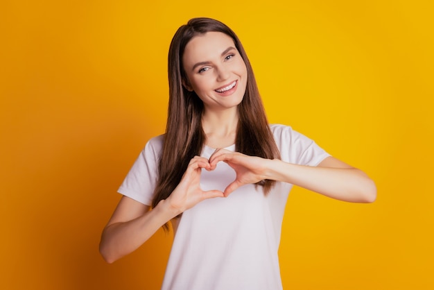 Photo d'une jolie dame mignonne montrant un signe de coeur portant un t-shirt blanc posant sur fond jaune