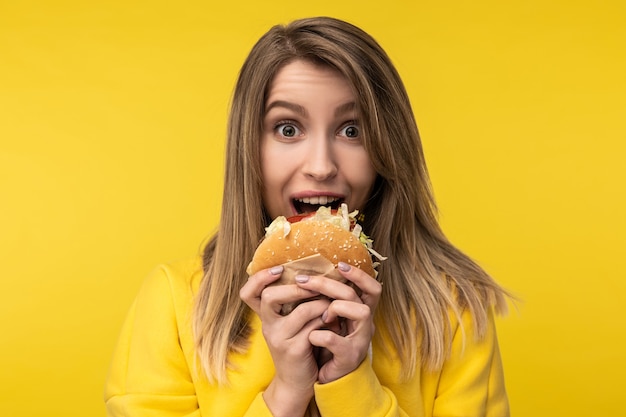 Photo de jolie dame pose d'une manière joyeuse tente de mordre un hamburger. Porte un sweat à capuche jaune décontracté, fond de couleur jaune isolé.