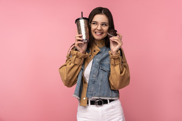 Photo de jolie dame tient du thé, du café et des muffins, attendez heureux quelque chose, portez une veste de jeans décontractée sous-chemise blanche isolée fond de couleur rose.