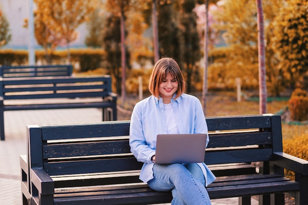 Photo d'une jolie étudiante assise sur un banc dans un parc verdoyant parcourant un ordinateur portable discutant avec des amis