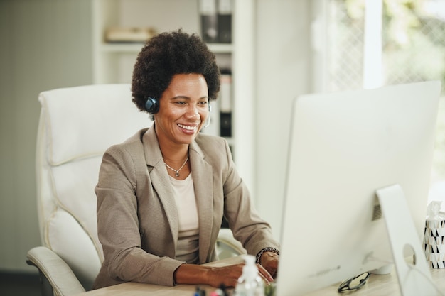 Photo d'une jolie femme d'affaires africaine assise seule dans son bureau à domicile avec des écouteurs et travaillant sur ordinateur pendant la pandémie de COVID-19.