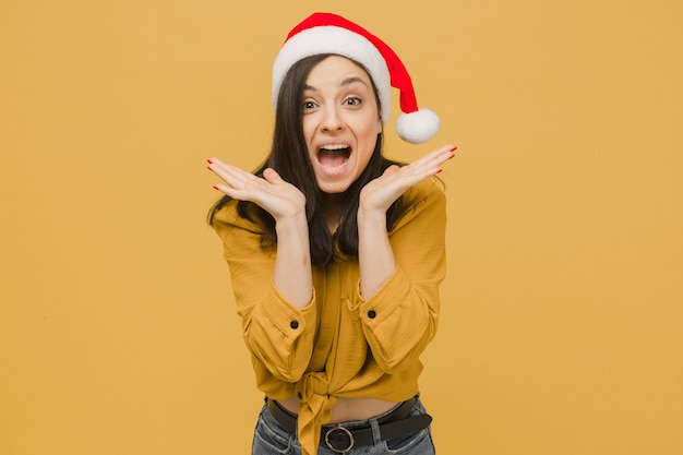 Photo la photo d'une jolie femme au chapeau de noël est surprise par les cadeaux. porte une chemise jaune, un fond de couleur jaune isolé.