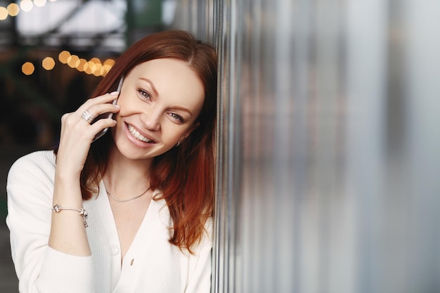 Photo d'une jolie femme aux cheveux roux avec un sourire positif a une conversation téléphonique avec des appels de clients à un ami vêtu d'une chemise blanche regarde la caméra avec une expression satisfaite aime la communication