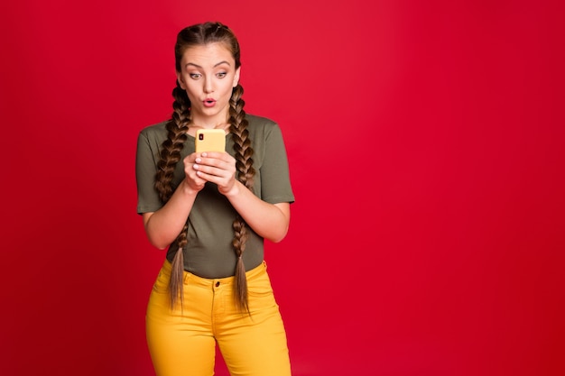 Photo de jolie femme drôle de longues tresses tenant le téléphone la lecture de nouveaux commentaires positifs bouche ouverte porter des pantalons jaunes décontractés t-shirt vert isolé fond de couleur rouge