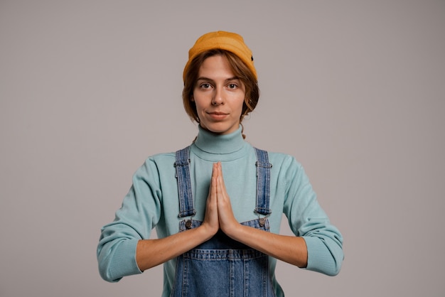 La photo de la jolie femme hipster montre se concentrer et se détendre. Femme blanche porte une combinaison en denim et un chapeau isolé sur fond de couleur grise.