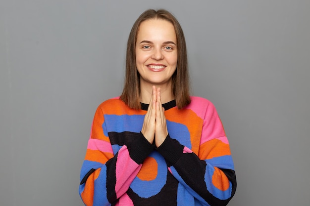 Photo d'une jolie femme positive souriante aux cheveux bruns portant un pull coloré posant isolé sur fond gris debout avec un geste de prière regardant la caméra avec un sourire à pleines dents