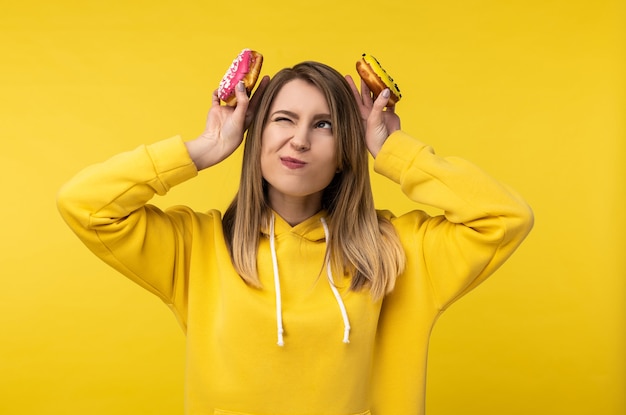 Photo d'une jolie femme tient des beignets derrière sa tête, faisant semblant d'être un lapin. Porte un sweat à capuche jaune décontracté, fond de couleur jaune isolé.