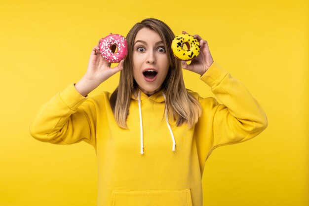 Photo d'une jolie femme tient des beignets devant ses yeux avec la bouche ouverte et le visage choqué. Porte un sweat à capuche jaune décontracté, fond de couleur jaune isolé.