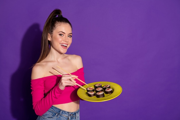 Photo d'une jolie fille étudiante tenir la plaque de baguettes manger des sushis asiatiques porter un haut rose recadré fond de couleur violet isolé