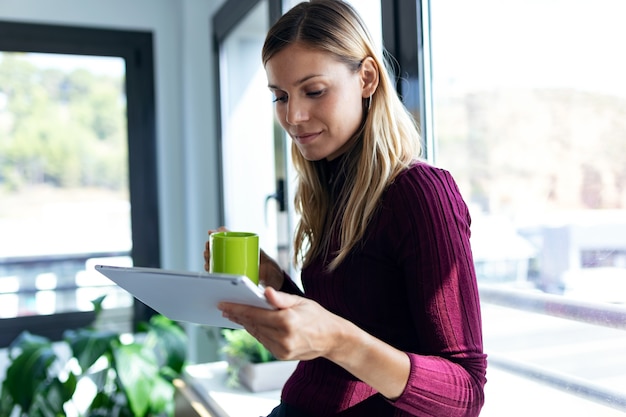 Photo d'une jolie jeune femme d'affaires utilisant sa tablette numérique et tenant une tasse de café assise à côté de la fenêtre du bureau.