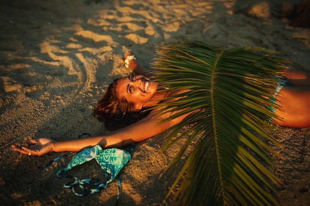 Photo d'une jolie jeune femme allongée sur la plage de sable tropicale recouverte de feuilles de palmier.