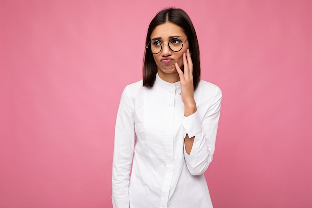 Photo d'une jolie jeune femme brune insatisfaite triste et bouleversée portant une chemise blanche et des lunettes optiques élégantes isolées sur fond rose regardant sur le côté.