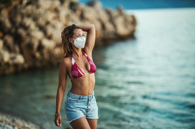 Photo d'une jolie jeune femme heureuse avec masque chirurgical profitant de vacances sur la plage pendant le COVID-19.