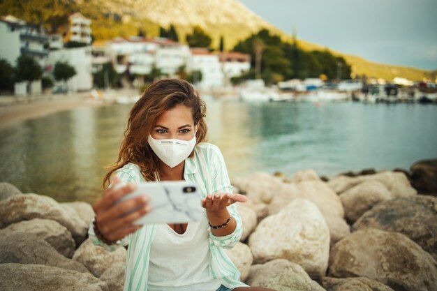 Photo d'une jolie jeune femme heureuse portant un masque de protection N95 et passant un appel vidéo avec son smartphone tout en passant du temps au bord de la mer pendant le COVID-19.