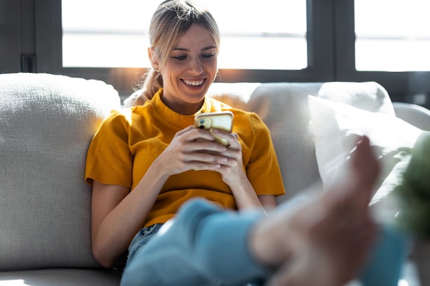 Photo d'une jolie jeune femme souriante utilisant un téléphone portable assis sur un canapé à la maison