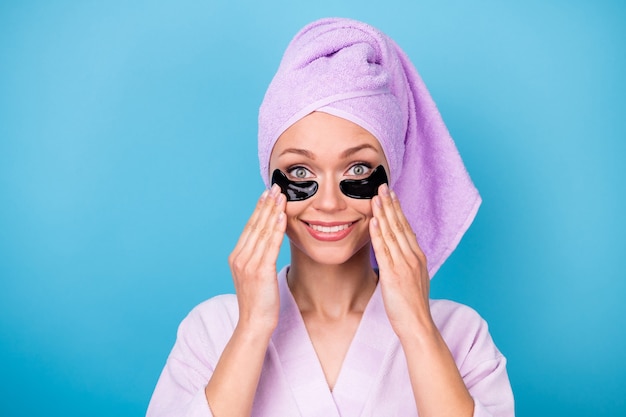 Photo d'une jolie jeune femme tenir des patchs pour les yeux porter une serviette violette turban peignoir de bain isolé sur fond de couleur bleu