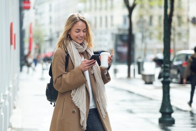 Photo d'une jolie jeune femme utilisant son téléphone portable tout en tenant une tasse de café dans la rue.