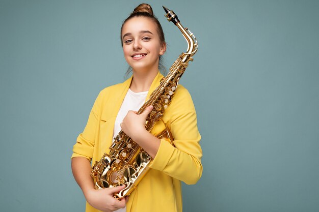 Photo d'une jolie petite fille brune souriante et positive portant une veste jaune à la mode, isolée sur un mur de fond bleu tenant un saxophone en regardant la caméra. Espace vide, espace de copie