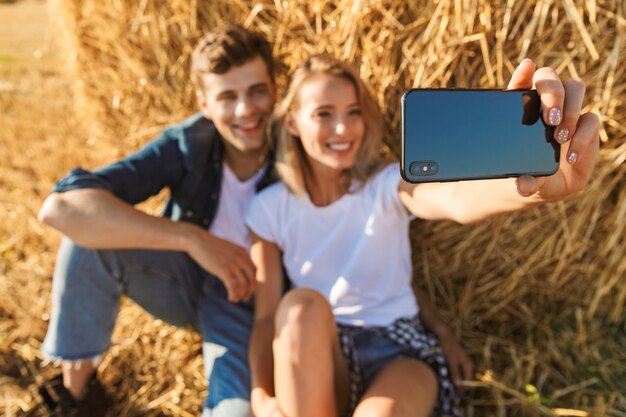 Photo de joyeux couple homme et femme prenant selfie sur smartphone alors qu'il était assis sous une grosse botte de foin dans le champ d'or, pendant la journée ensoleillée