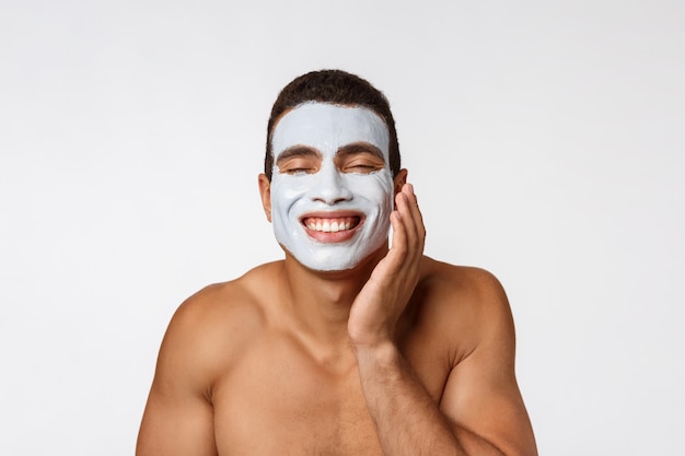 Photo de joyeux gentil jeune homme souriant à la caméra. Portrait d'un homme avec un masque crème sur les joues.