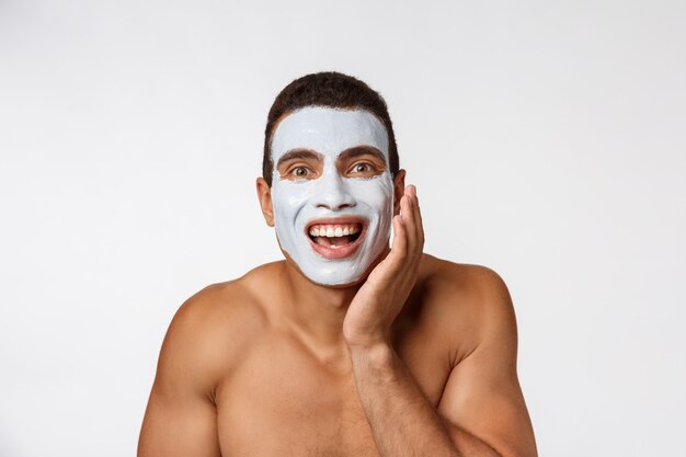 Photo de joyeux gentil jeune homme souriant à la caméra. Portrait d'un homme avec un masque crème sur les joues.