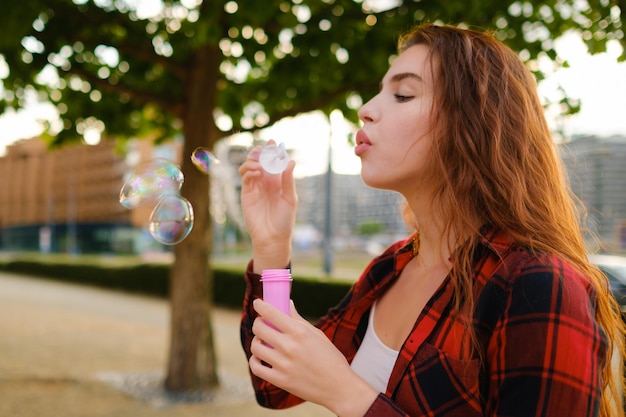 Photo latérale de profil d'une jeune femme portant une chemise à carreaux tendance soufflant des bulles de savon dans la rue