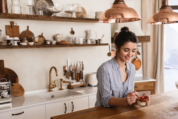 Photo d'une magnifique femme brune mangeant un dessert à la panna cotta avec une cuillère à café tout en prenant son petit-déjeuner dans une cuisine élégante à la maison