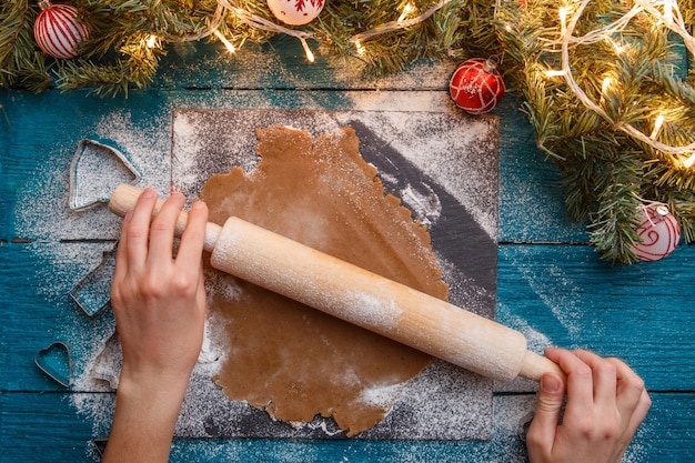 Photo de mains humaines, rouleau à pâtisserie, pâte, branches d'épinette, guirlandes sur table bleue