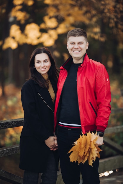 Photo de maman aux longs cheveux noirs en manteau noir, joli papa aux cheveux courts et noirs en veste rouge tenir le bouquet de feuilles d'automne