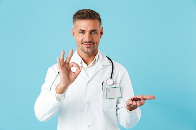 Photo d'un médecin de bel homme mûr posant isolé sur mur de mur bleu tenant des vitamines de pilules montrant le geste correct.