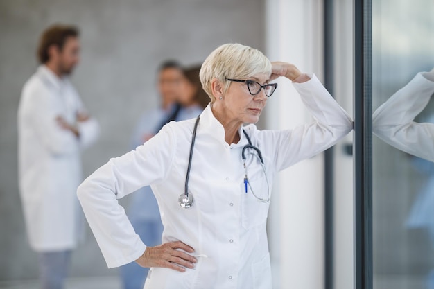 Photo d'un médecin senior qui a l'air inquiet en se tenant debout et pensif en regardant par la fenêtre dans un couloir d'hôpital pendant la pandémie de Covid-19.