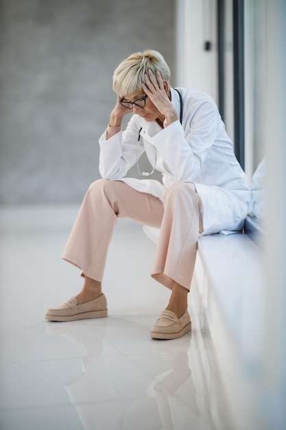 Photo d'un médecin senior qui a l'air stressé assis sur le sol près d'une fenêtre dans un couloir d'hôpital vide pendant la pandémie de Covid-19.