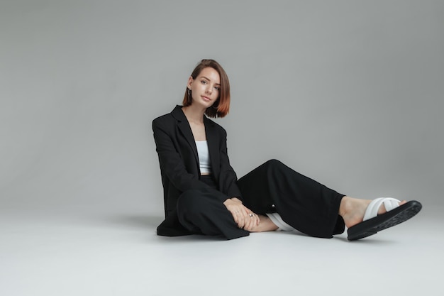 Une photo de mode d'une femme à la mode en costume noir assise sur le sol dans un studio