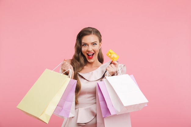 Photo de mode riche femme des années 20 en robe souriant tout en tenant différents sacs à provisions et carte de crédit en mains, isolé sur mur rose