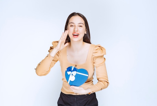 Photo d'un modèle de jeune femme tenant une boîte-cadeau en forme de coeur contre un mur blanc.