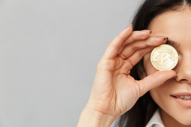 Photo de moitié côté d'une femme riche aux longs cheveux bruns couvrant ses yeux avec du bitcoin doré, isolé sur mur gris